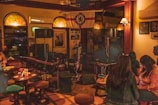 A dimly lit bar setting with several people sitting at tables and a small stage area with a microphone. Wall decorations include a Chelsea football club flag and framed photographs. The atmosphere suggests a casual, relaxed gathering.