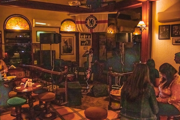A dimly lit bar setting with several people sitting at tables and a small stage area with a microphone. Wall decorations include a Chelsea football club flag and framed photographs. The atmosphere suggests a casual, relaxed gathering.