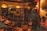 A dimly lit bar setting with several people sitting at tables and a small stage area with a microphone. Wall decorations include a Chelsea football club flag and framed photographs. The atmosphere suggests a casual, relaxed gathering.