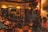 A dimly lit bar setting with several people sitting at tables and a small stage area with a microphone. Wall decorations include a Chelsea football club flag and framed photographs. The atmosphere suggests a casual, relaxed gathering.