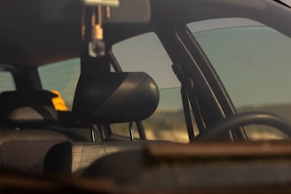 The interior of a car is viewed through a window with a focus on the dark leather headrest. There is a yellow object on the back seat, and the window reflects some daylight, giving a soft, warm tone to the scene.