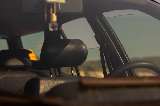 The interior of a car is viewed through a window with a focus on the dark leather headrest. There is a yellow object on the back seat, and the window reflects some daylight, giving a soft, warm tone to the scene.