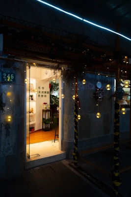 A warmly lit entrance leads into a cozy, inviting space decorated with shelves and plants. String lights hang outside the doorway, casting a gentle glow against the dark exterior.