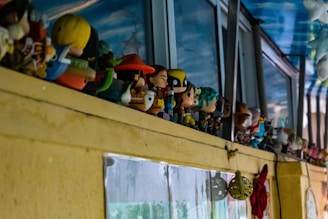 A collection of various figurines is lined up on a yellow shelf against a backdrop of windows and a blue sky ceiling. The figurines are diverse, featuring colorful characters, some with distinctive hats and outfits. Below the shelf, part of a wall with pictures and a red ribbon or cloth hanging is visible.