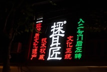 Neon sign with Chinese characters in vibrant colors. The sign includes large white characters in the center, surrounded by red and green text on a dark background, suggesting a nighttime setting. The characters seem to be advertising or indicating direction.