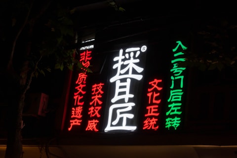 Neon sign with Chinese characters in vibrant colors. The sign includes large white characters in the center, surrounded by red and green text on a dark background, suggesting a nighttime setting. The characters seem to be advertising or indicating direction.