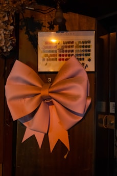 A large, pink bow is prominently displayed against a dimly lit wooden surface. Above the bow, there is a framed board with multicolored palettes, possibly samples or designs. The ambient lighting casts warm shadows, and there are floral decorations partially visible on the left side.