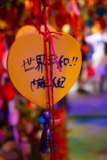 A heart-shaped yellow plaque with handwritten text in Chinese hangs by a red string, with a small bell attached. The background is out of focus with vibrant colors, creating a festive and lively atmosphere.