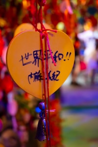 A heart-shaped yellow plaque with handwritten text in Chinese hangs by a red string, with a small bell attached. The background is out of focus with vibrant colors, creating a festive and lively atmosphere.