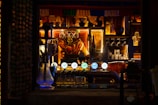 A dimly lit bar with a variety of glassware and decorations on shelves behind a counter. There are several beer taps with illuminated labels, glasses hanging upside down, and various bottles and ornaments arranged on the shelves. The scene is warm and cozy with ambient lighting.