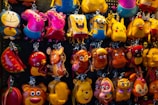 A colorful display of various character keychains hanging on a pegboard. The keychains include popular animated characters such as a yellow sponge with square pants, a grinning cat, a yellow dinosaur, and pink animals with snouts. They are made from materials that give them a shiny, glossy look, and the background is formed by a dark pegboard with small holes.