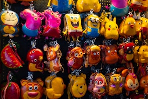 A colorful display of various character keychains hanging on a pegboard. The keychains include popular animated characters such as a yellow sponge with square pants, a grinning cat, a yellow dinosaur, and pink animals with snouts. They are made from materials that give them a shiny, glossy look, and the background is formed by a dark pegboard with small holes.