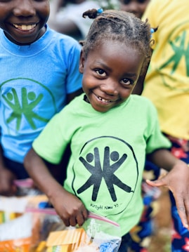 A smiling child in a green t-shirt holding a plastic bag filled with snacks stands in the foreground, with another child in a blue t-shirt and more children in the background. The children are wearing shirts with similar logos consisting of a circle with three figures inside.