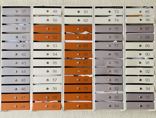 A grid of numbered mailboxes arranged in several columns, each containing multiple slots. Some of the mailboxes have letters and papers sticking out of them. The mailboxes are colored in shades of white, grey, and orange, and each slot is labeled with a different number.