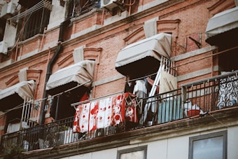 A brick building with multiple windows, each covered with a small beige awning. Clothes and towels are hanging on a railing outside, suggesting a laundry drying area. There are various items and clotheslines attached to the railing.