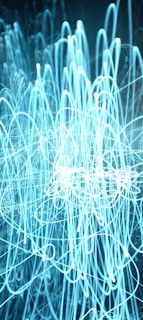 A chaotic mesh of light trails forming intricate loops and overlapping patterns against a dark background. The lines are predominantly blue and white, suggesting motion and a dynamic energy.
