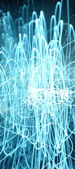 A chaotic mesh of light trails forming intricate loops and overlapping patterns against a dark background. The lines are predominantly blue and white, suggesting motion and a dynamic energy.