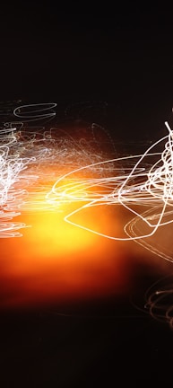 An abstract display featuring swirling lines of light in white and orange hues against a dark background, creating a sense of motion and energy.