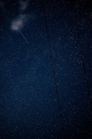 A starry night sky with a faint shooting star visible among the stars. Wisps of clouds are present in the upper left corner. A vertical dark line, resembling a wire or cable, cuts through the sky.
