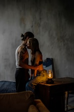 A dimly lit room where a couple is sharing an intimate moment. The woman is sitting on a wooden surface next to a small lamp, with subtle light highlighting her face and the man's arms. The man stands close, partially facing away, with his arm around her waist. A quiet, moody atmosphere is set by the textured grey wall and minimalistic furnishings, including a sofa and a piece of abstract art leaning against the wall.