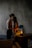 A dimly lit room where a couple is sharing an intimate moment. The woman is sitting on a wooden surface next to a small lamp, with subtle light highlighting her face and the man's arms. The man stands close, partially facing away, with his arm around her waist. A quiet, moody atmosphere is set by the textured grey wall and minimalistic furnishings, including a sofa and a piece of abstract art leaning against the wall.