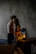 A dimly lit room where a couple is sharing an intimate moment. The woman is sitting on a wooden surface next to a small lamp, with subtle light highlighting her face and the man's arms. The man stands close, partially facing away, with his arm around her waist. A quiet, moody atmosphere is set by the textured grey wall and minimalistic furnishings, including a sofa and a piece of abstract art leaning against the wall.