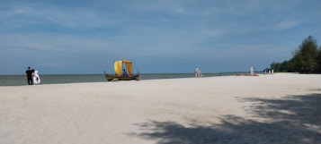A tranquil beach scene features a wide expanse of white sand leading to calm waters under a clear blue sky. A traditional wooden boat with a yellow canopy sits on the sand, while a couple strolls along the shore. Sparse trees and distant beach amenities can be seen in the background.