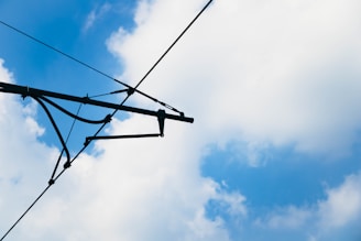 A section of electrical power lines for trains is visible against a backdrop of a partly cloudy sky. The cables are black and intersect at several points, creating a geometric pattern.