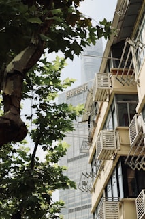 A tall building in the background with the word 'Regent' visible, partially obscured by trees. In the foreground, there is a residential building with multiple balconies, air conditioning units, and a clear view of the windows. The scene is interspersed with green leaves from trees, creating a contrast between nature and urban architecture.