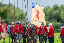A group of people dressed in historical military attire, featuring red coats and metal helmets, stand in a grassy field. They are holding long wooden pikes and a large flag with a red lion emblem and the words 'God Save the King' written on it. The background includes trees and a cloudy sky, suggesting an outdoor reenactment or event.