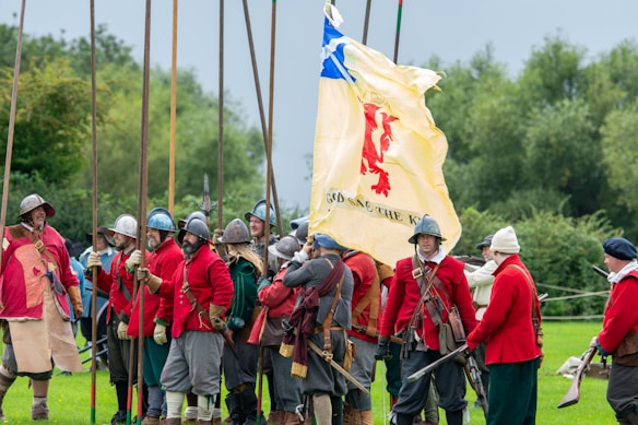 A group of people dressed in historical military attire, featuring red coats and metal helmets, stand in a grassy field. They are holding long wooden pikes and a large flag with a red lion emblem and the words 'God Save the King' written on it. The background includes trees and a cloudy sky, suggesting an outdoor reenactment or event.