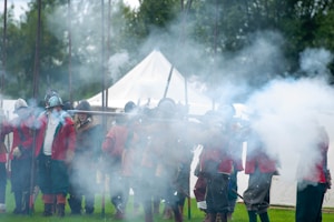 A group of reenactors dressed in historical military uniforms are engaging in a historical reenactment. They are wearing helmets and red coats, and are surrounded by smoke, suggesting the discharge of muskets. The setting appears to be outdoors, with green grass and tents in the background. The scene is lively with a sense of action and movement.