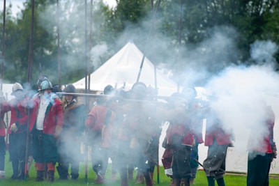A group of reenactors dressed in historical military uniforms are engaging in a historical reenactment. They are wearing helmets and red coats, and are surrounded by smoke, suggesting the discharge of muskets. The setting appears to be outdoors, with green grass and tents in the background. The scene is lively with a sense of action and movement.