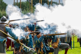 A group of historical reenactors in traditional military uniforms are firing muskets, creating large plumes of white smoke. They wear helmets and blue outfits with leather accessories, positioned on a grassy field.