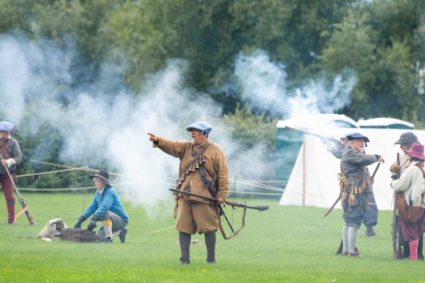 A group of people dressed in historical clothing engage in a re-enactment, with smoke surrounding them, possibly from musket fire. They are positioned on a grassy field with tents in the background, suggesting a camp setting.
