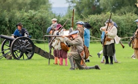 A reenactment scene depicting a group of individuals dressed in historical attire, engaging in a mock battle. One person is kneeling and firing a musket, while others stand behind with various weapons. A vintage cannon is positioned to the side. The participants are on a grassy field surrounded by trees, suggesting an open outdoor location.