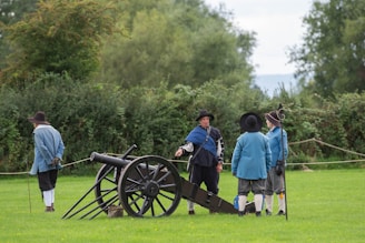 Several people dressed in historical clothing are gathered on a grassy field. A large, old-style cannon is positioned nearby, and the individuals appear to be engaged in conversation.