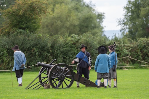 Several people dressed in historical clothing are gathered on a grassy field. A large, old-style cannon is positioned nearby, and the individuals appear to be engaged in conversation.