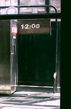 A metallic structure with a digital display showing the time '12:00'. The environment appears industrial, with shadows and sunlight casting patterns on the floor. A red and white sticker is affixed to the metal surface.