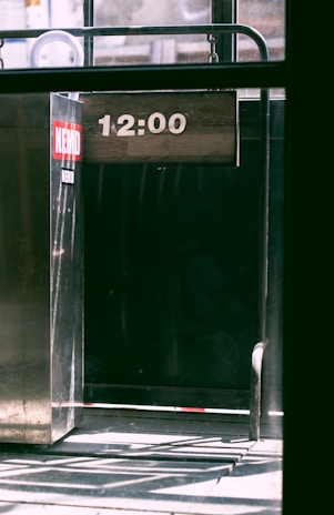 A metallic structure with a digital display showing the time '12:00'. The environment appears industrial, with shadows and sunlight casting patterns on the floor. A red and white sticker is affixed to the metal surface.