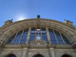 An ornate architectural structure with a large arched facade featuring detailed sculptures and decorative elements. A clock is centrally positioned above the archway. The building is adorned with classical columns and stone carvings, and a sign reads 'Hauptbahnhof' along with the DB logo.