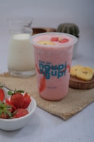 A pink smoothie drink is surrounded by fresh strawberries in a bowl, a bottle of milk, and a small cactus plant. The drink is topped with banana and strawberry slices and is placed on a textured fabric, possibly burlap.