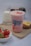 A pink smoothie drink is surrounded by fresh strawberries in a bowl, a bottle of milk, and a small cactus plant. The drink is topped with banana and strawberry slices and is placed on a textured fabric, possibly burlap.