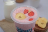 A creamy pink smoothie topped with banana and strawberry slices in a clear plastic cup. In the background, there's a glass bottle of milk and a few banana slices on a wooden board.