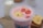 A creamy pink smoothie topped with banana and strawberry slices in a clear plastic cup. In the background, there's a glass bottle of milk and a few banana slices on a wooden board.