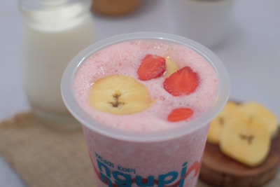 A creamy pink smoothie topped with banana and strawberry slices in a clear plastic cup. In the background, there's a glass bottle of milk and a few banana slices on a wooden board.