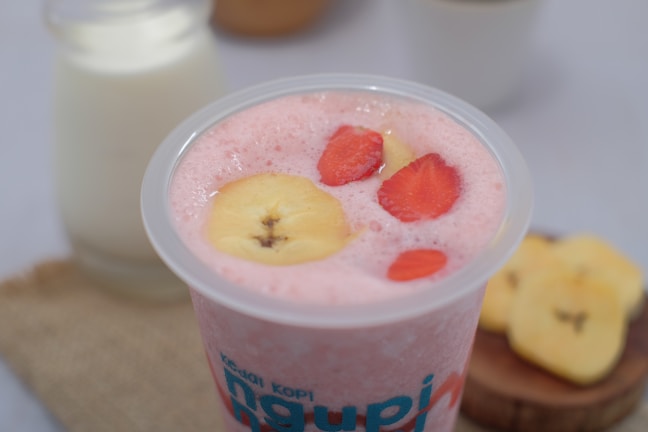 A creamy pink smoothie topped with banana and strawberry slices in a clear plastic cup. In the background, there's a glass bottle of milk and a few banana slices on a wooden board.