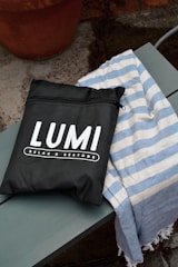 A black bag with white lettering displaying the brand name 'LUMI' and the phrase 'RELAX & RESTORE' is placed on a wooden bench. Next to the bag is a blue and white striped fabric, possibly a towel. The background features a concrete surface with a terracotta pot.