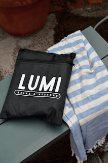 A black bag with white lettering displaying the brand name 'LUMI' and the phrase 'RELAX & RESTORE' is placed on a wooden bench. Next to the bag is a blue and white striped fabric, possibly a towel. The background features a concrete surface with a terracotta pot.