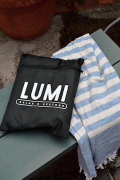 A black bag with white lettering displaying the brand name 'LUMI' and the phrase 'RELAX & RESTORE' is placed on a wooden bench. Next to the bag is a blue and white striped fabric, possibly a towel. The background features a concrete surface with a terracotta pot.